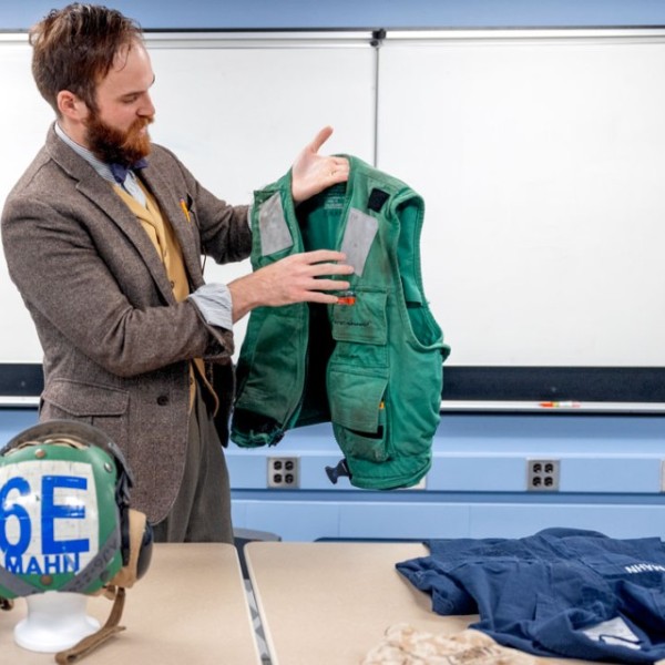 A man, Christopher Mahn, holds up a float coat he wore as a mechanic on an aircraft carrier in a classroom. Other military items litter the table in front of him.