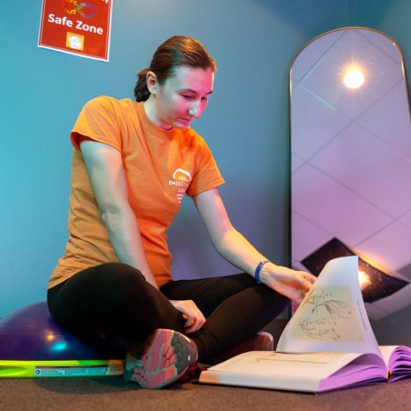 A student sits crosslegged on a half exercise ball reading from a book.