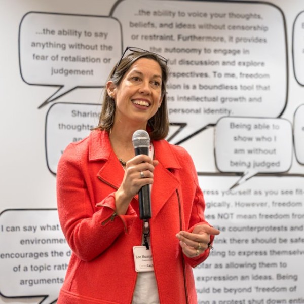 A woman in a bright red blazer speaks into a microphone in front of a wall covered with speech bubbles.