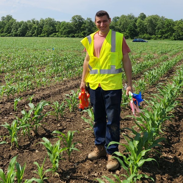 Sadiel standing in a farm field wearing a fluorescent vest and carrying research equipment.