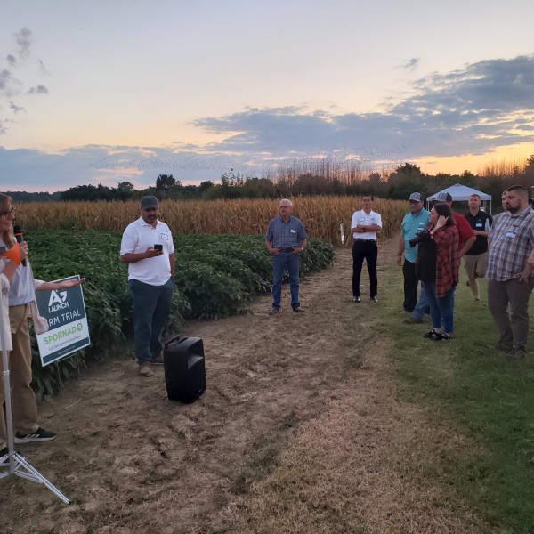 A small group of ten researchers listen to a speaker outside on the edge of a crop field.