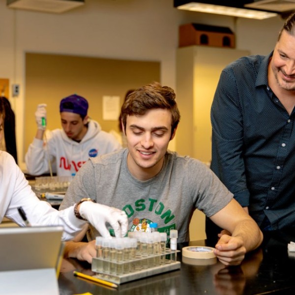 A female and male student work with a male teacher in a laboratory classroom holding vials. 