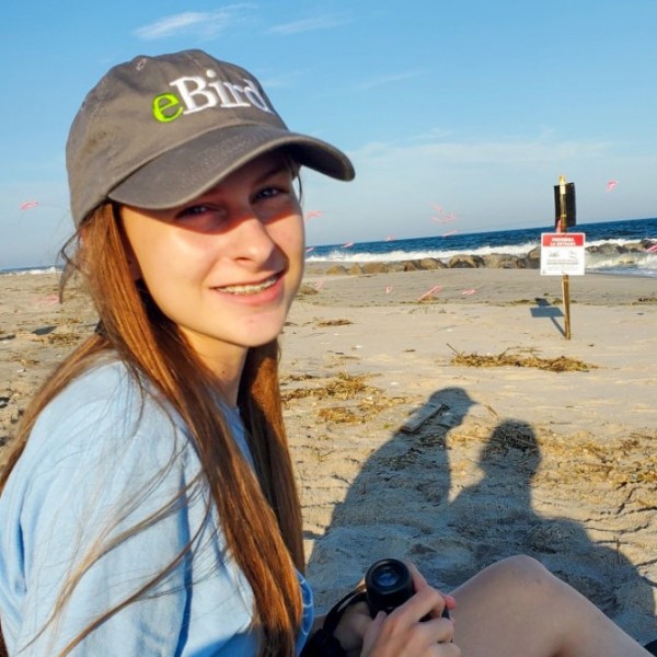 A girl in a hat holding binoculars sits on a beach.