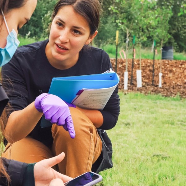 A woman wearing purple gloves and holding a blue folder crouches in a field and speaks to another woman in a mask.