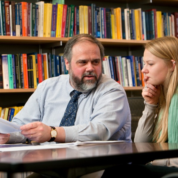 An older man speaks with a female student in front of a bookcase.