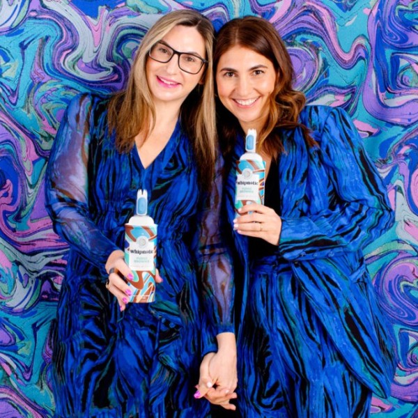 Two women hold cans of whipped cream, labeled "whipnotic", in front of a blue swirly background.