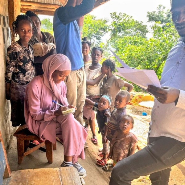 A black woman in a baby pink modest outfit and head covering sits and takes notes on an pad. A black male researcher stands and holds a piece of paper. They are surrounded by nine family members speaking.