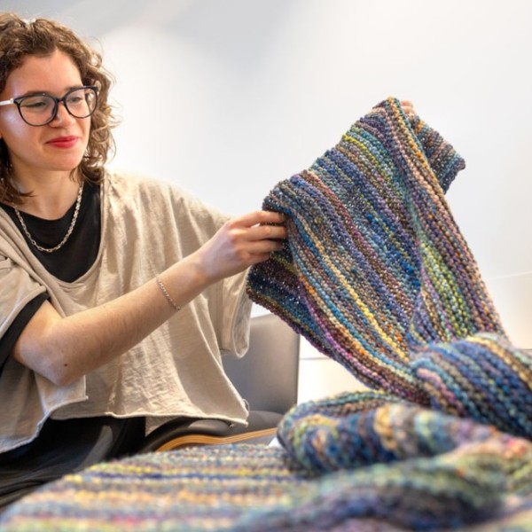 A woman holds up a long rainbow knitted blanket.