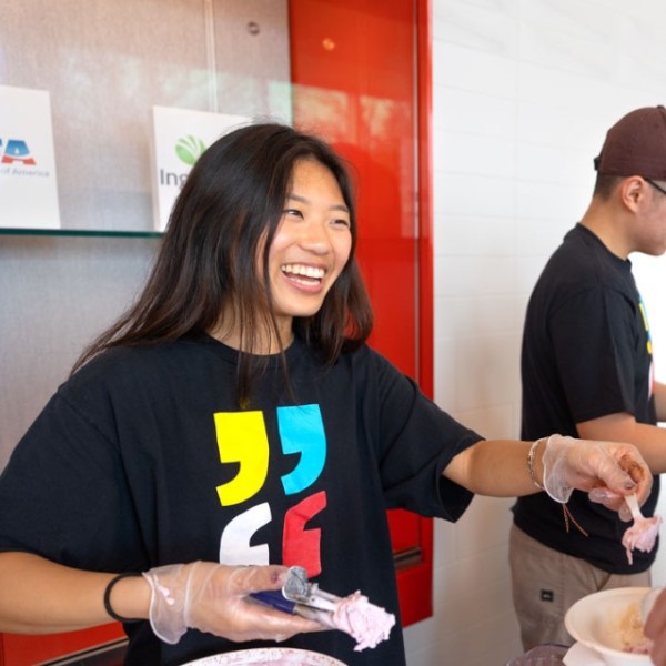 A woman with long brown hair, wearing a freedom of expression year shirt with four multicolored quotation mark, scoops ice cream into a mans cup.