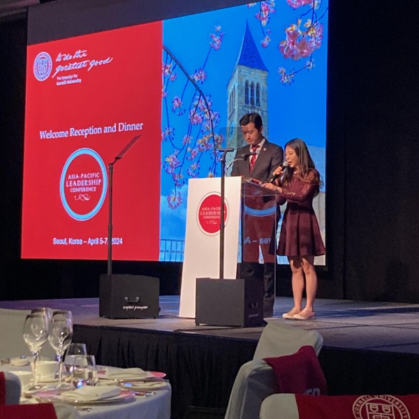 stage with man and woman standing in front of screen speaking into microphone