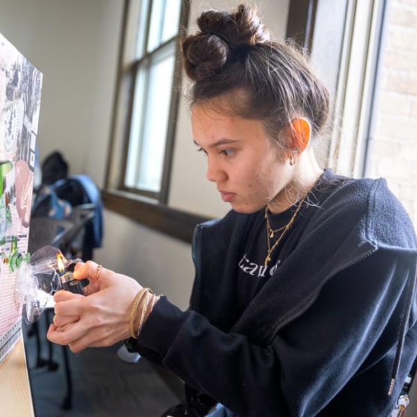 Kailee Tomas ’26, an environment and sustainability major in the College of Agriculture and Life Sciences, uses a lighter to melt plastic litter collected from the Mohawk River watershed. The litter is attached to the canvases in the shape of the river, over a collage of newspaper articles about the Haudenosaunee people past and present.