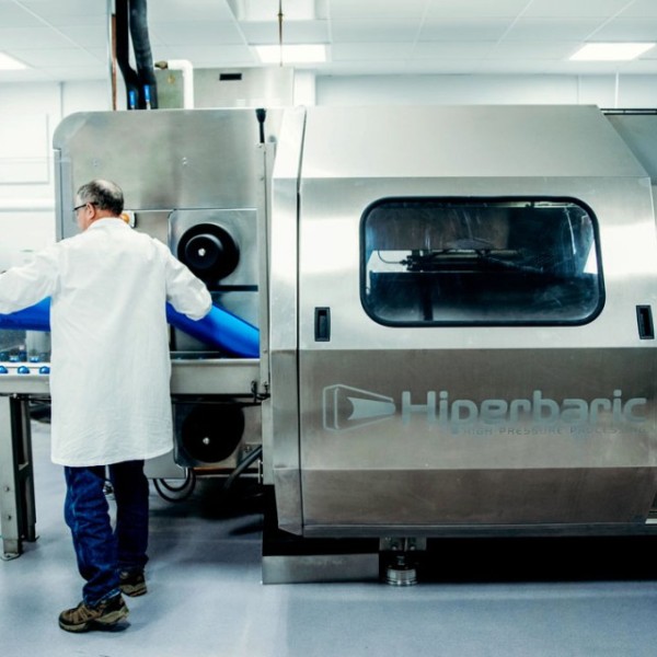 A man in a lab coat loads a blue tube into a metal chamber labeled "Hiperbaric High Pressure Processing."