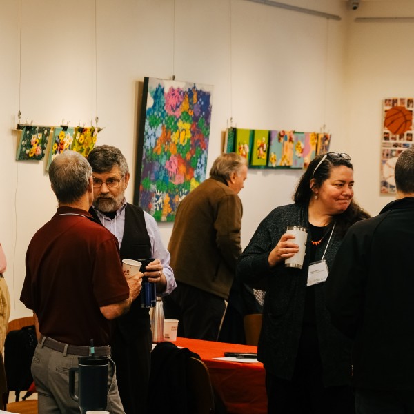 Photograph of a small group of people talking with coffee cups in hand with some art hanging behind them on the walls.