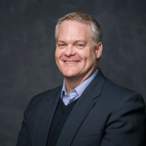 Headshot of Christopher Barrett, in a black suit infront of a grey background.
