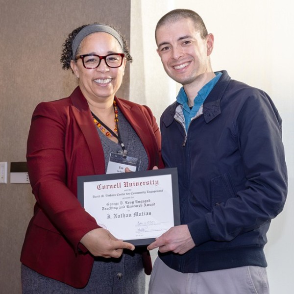 A woman in a red blazer, brown curly hair, and glasses holds a piece of paper with a man with a buzz cut and a blue jacket. It reads "George D. Levy Engaged Teaching and Research Award, J. Nathan Matais."