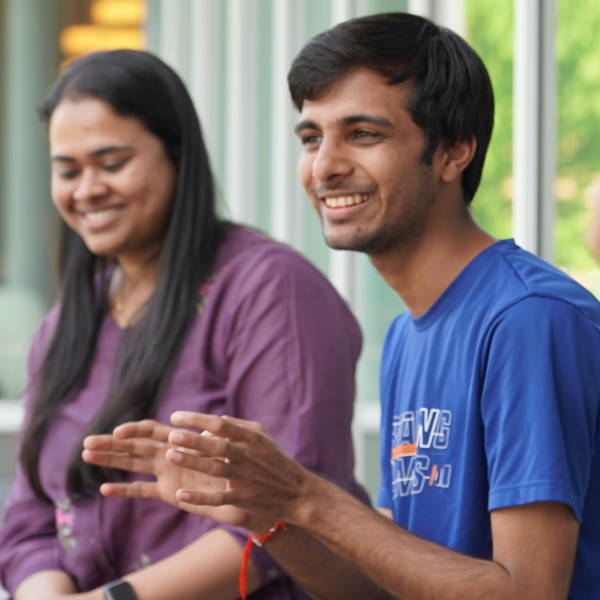 A woman is smiling on the left as a man on the right smiles and talks, gesturing with his hands.
