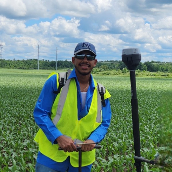 Man in a neon work vest smiling at the camera holding equipment in a field.
