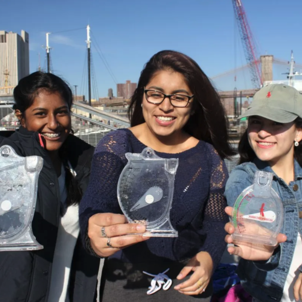 Photograph of three young women smiling holding clear scientific water gauges with a harbor in the background.