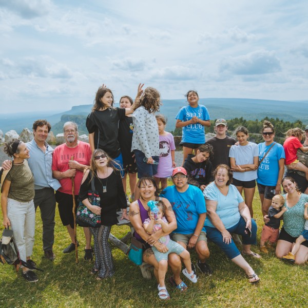 Photograph of a group of people from a variety of age groups standing on a grassy hill with rolling hills far into the background.