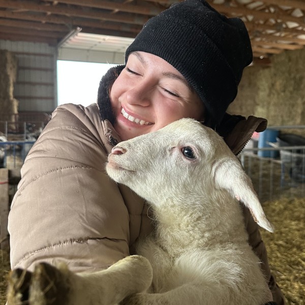 A woman wearing a hat hugging a baby animal in a barn.