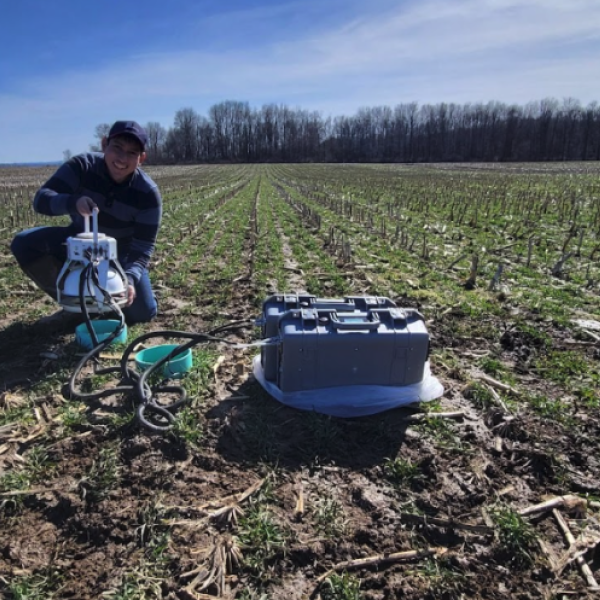 A man smiles while holding a LICOR greenhouse gas sampling system in a field.