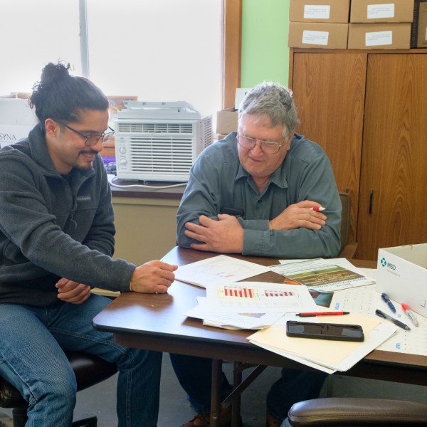 Two men sitting at a desk with papers on it talking.