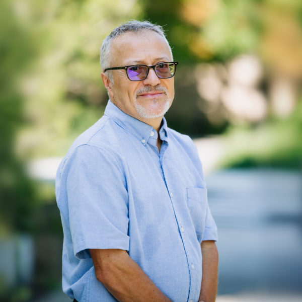 Toni stands in a shaded courtyard wearing a blue button-down shirt and glasses.