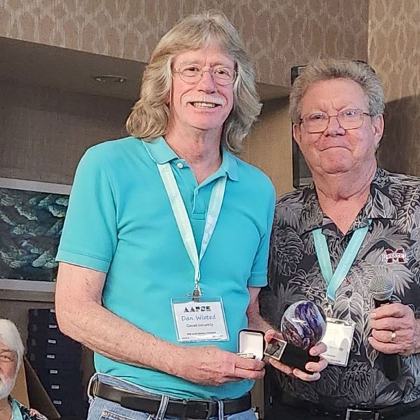 Two people standing next to each other wearing name badges and an award, smiling for he camera. There is another person sitting behind them in the background 