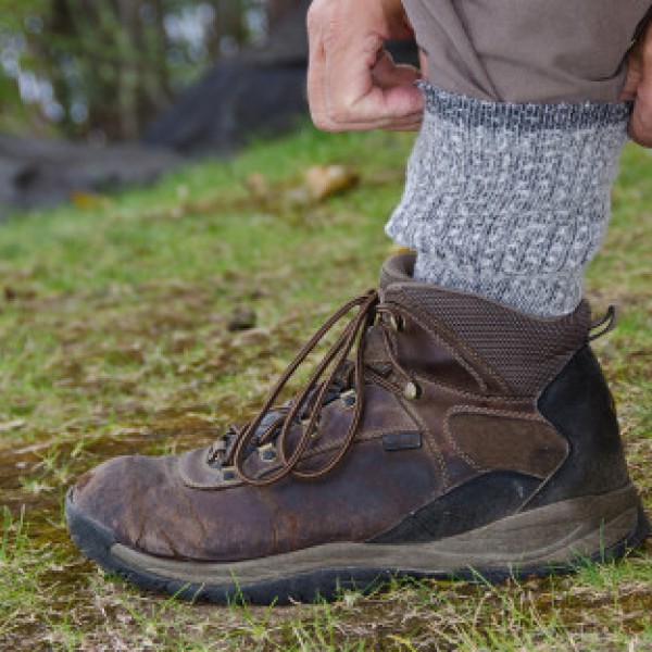 A close-up of a person outdoors adjusting their gray wool sock above a brown hiking boot on grassy ground.
