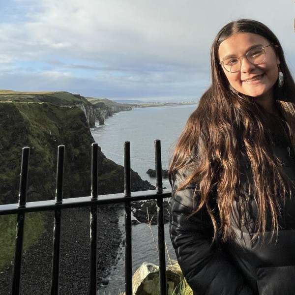 Smiling student in a black jacket stands by a railing overlooking dramatic coastal cliffs and the ocean.